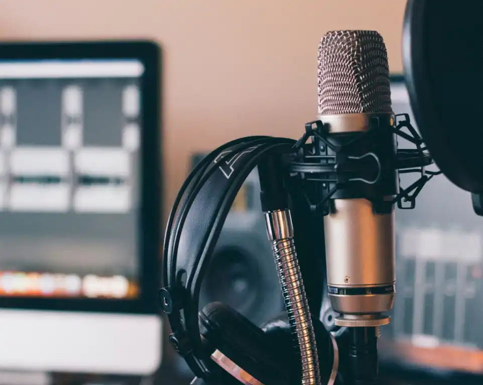 An immersive snapshot of podcasting: A sleek silver condenser microphone takes center stage in a spider stand, with a hint of a noise filter attached. Adjacent, headphones dangle, signaling a recent recording session. The frame expands to reveal a computer screen on the left, showcasing an audio-processing program in action. Audio waves on the screen signify meticulous editing, capturing the essence of the podcast creation process.