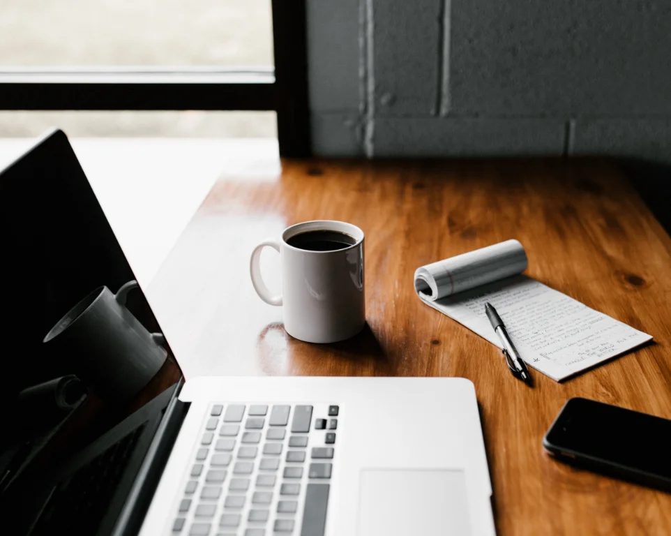 Desk setup with a laptop, coffee cup, notebook, pen, and phone.